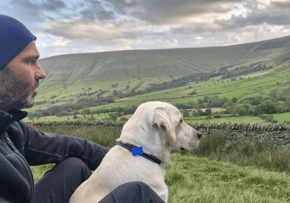Woodland Labrador Academy trainer sitting on a hillside beside a yellow Labrador, both looking out across green fells and drystone walls