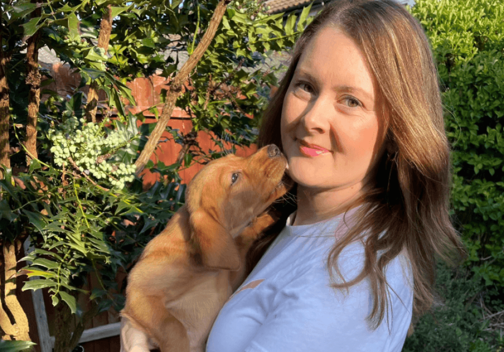 Woman holding a fox-red Labrador puppy in a sunny garden, the puppy nuzzling towards her